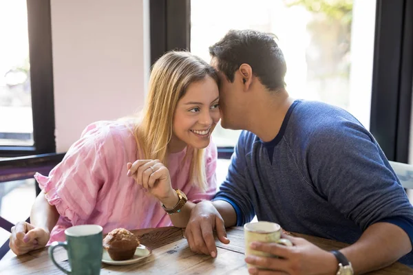 Young man whispering in a woman\'s ear while drinking coffee and having fun at a restaurant