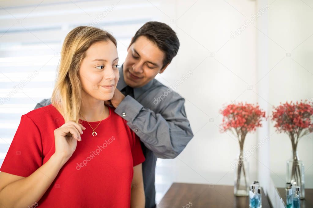 Loving young husband fastening necklace on woman's neck at home