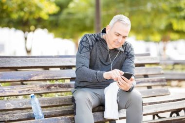 Bankta oturan ve açık havada bir parkta egzersiz sonra ile cep telefonu müzik dinlerken mutlu orta yaşlı adam