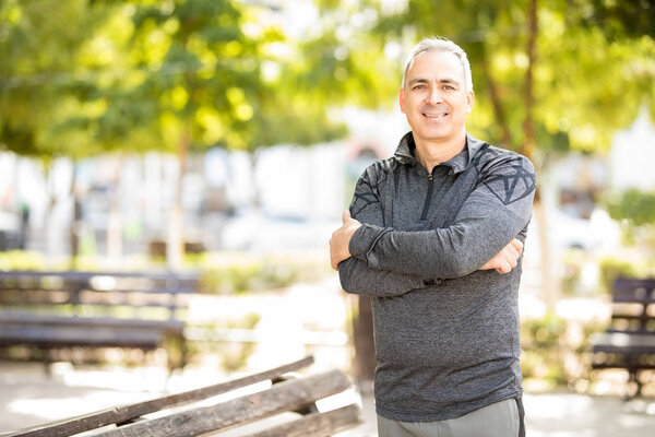 Portrait of healthy middle aged hispanic man standing outdoors in park with his arms crossed and smiling