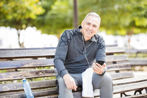 格好の成熟の肖像男は公園で屋外で運動から休憩をとりながらスマート フォンで音楽を聴く — ストック写真