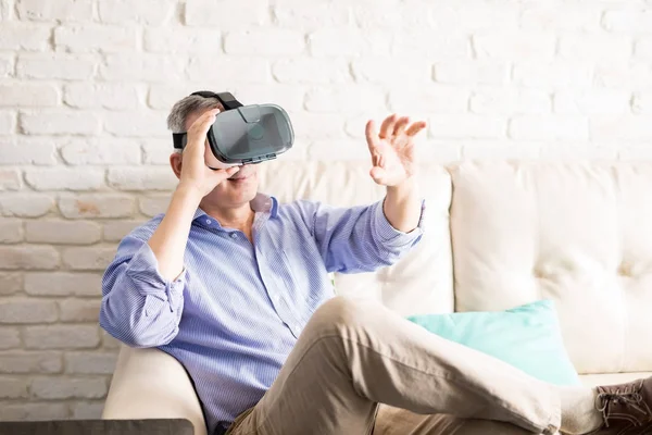 Man with gray hair using VR glasses and extending his hand forward to touch the graphics while sitting at home