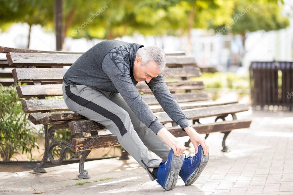 Mature Latin man sitting on a city park bench stretching and touching his toes