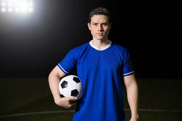 英俊的年轻足球运动员的肖像 当站在蓝色的制服 手持一个球在足球场上进行眼神接触 — 图库照片