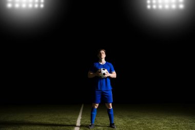 Mavi üniforma ayakta futbol üzerinde giyen futbolcu tam uzunlukta zemin bir top tutan ve bir göz teması yapma