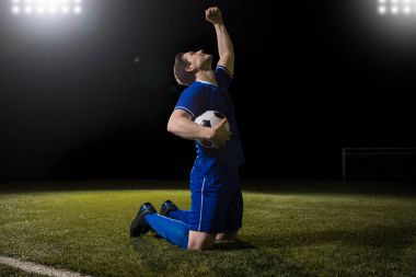 Excited young soccer player on knees and raising his arm after scoring a goal in match clipart