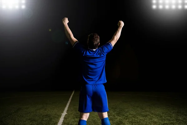 年轻橄榄球运动员的后方看法在蓝色制服站立用手胳膊在赢取比赛以后上升了 — 图库照片