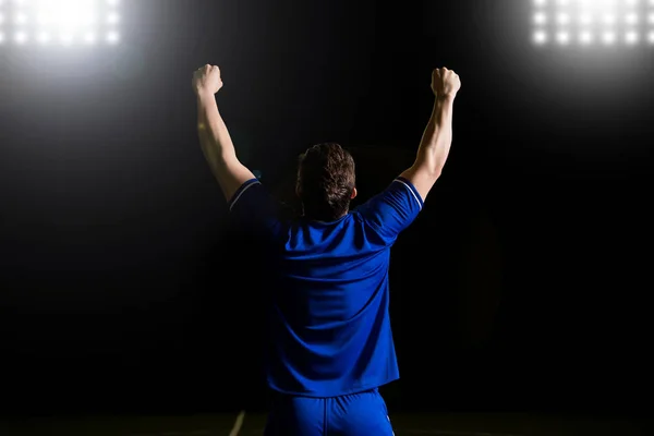 Rear view of soccer player in blue jersey with hands raised after scoring a goal in football match