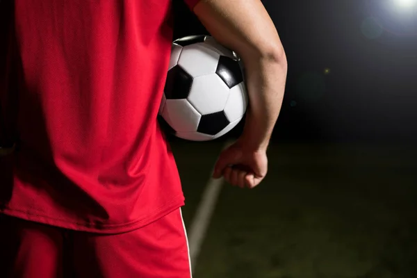 裁剪后视图的一个年轻的足球运动员在红色制服举行球在他的怀里在体育场 — 图库照片