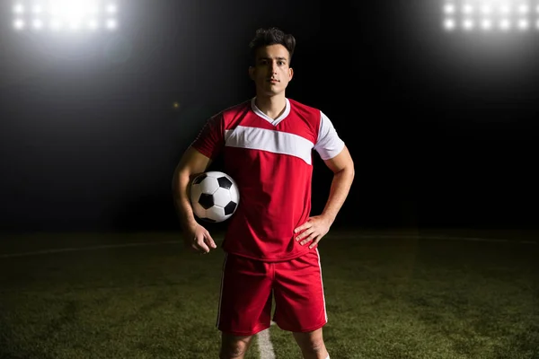 Jogador Futebol Vestindo Uniforme Vermelho Campo Futebol Sob Luzes Segurando — Fotografia de Stock