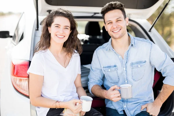 couple sitting with coffee in car trunk during break on road