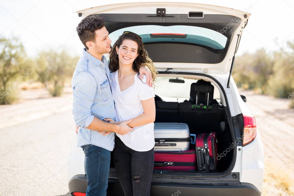 man and woman smiling and standing near car with luggage in trunk