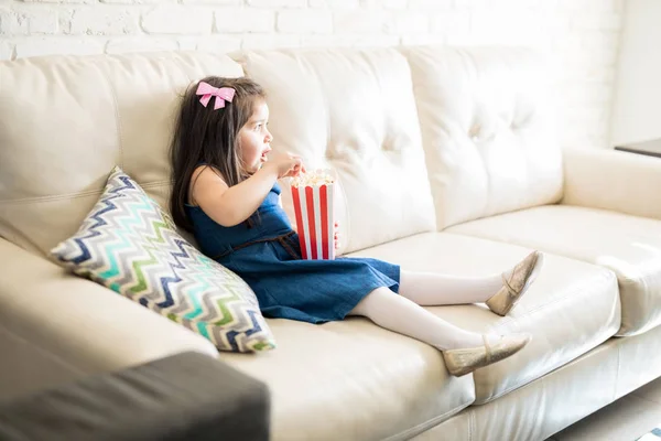 ポップコーンを食べたり 自宅のテレビで映画を見ているソファーに座っている少女 — ストック写真