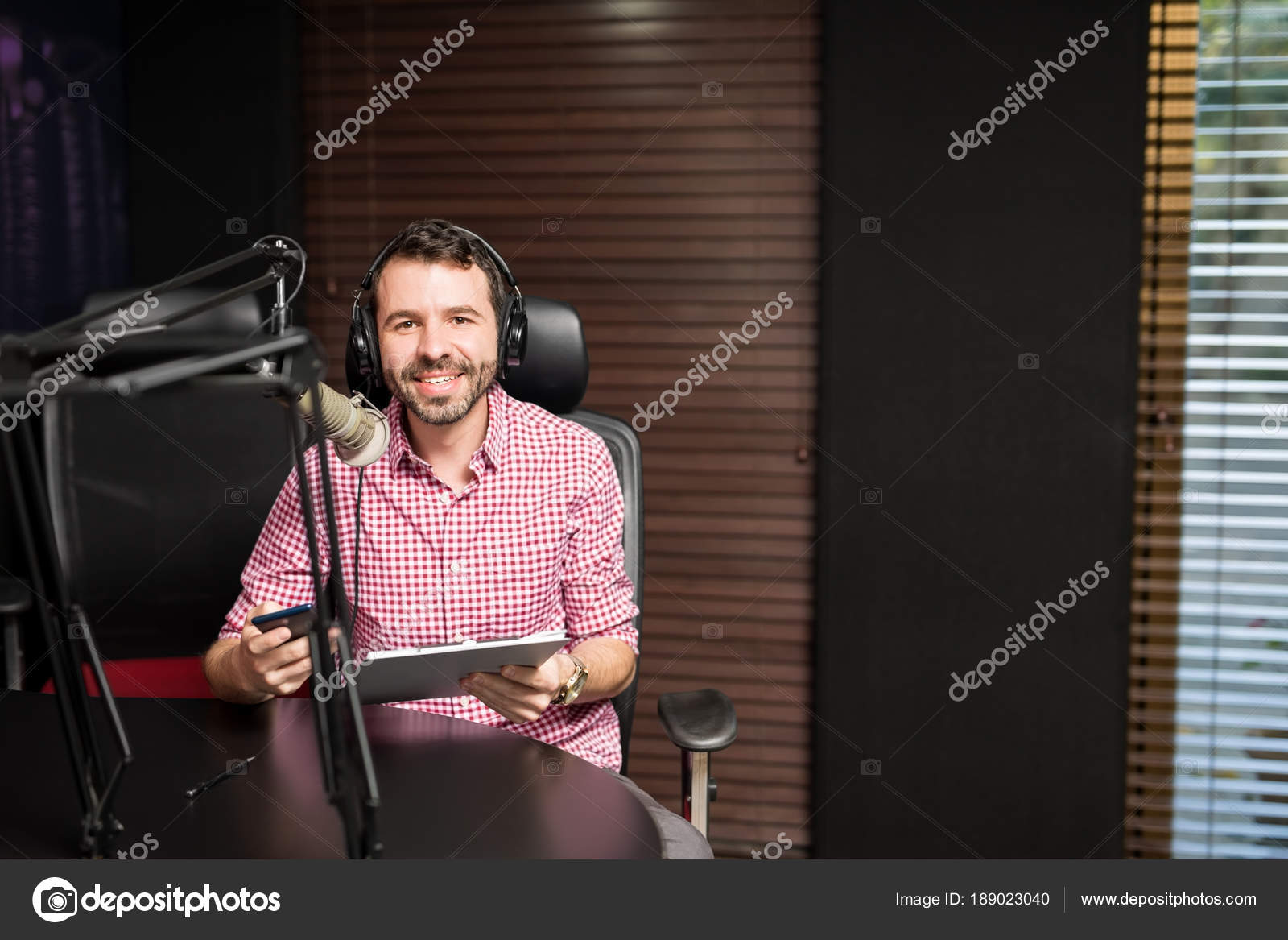 Young Latin Man Sitting Microphone Holding Clipboard Using Smartphone