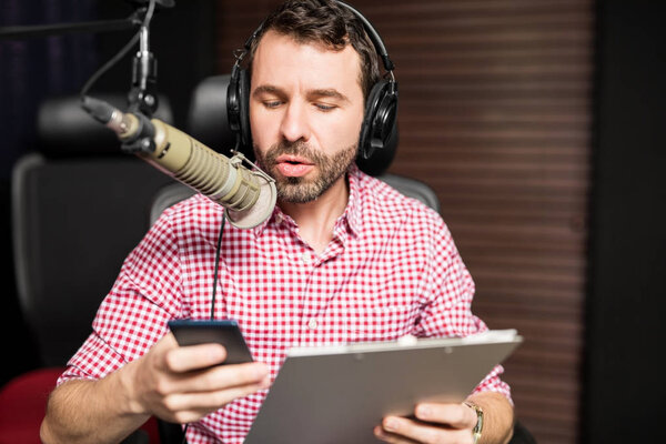 young latin man sitting near microphone holding clipboard and using smartphone at radio station