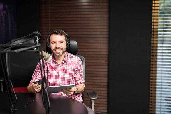 young latin man sitting near microphone holding clipboard and using smartphone at radio station