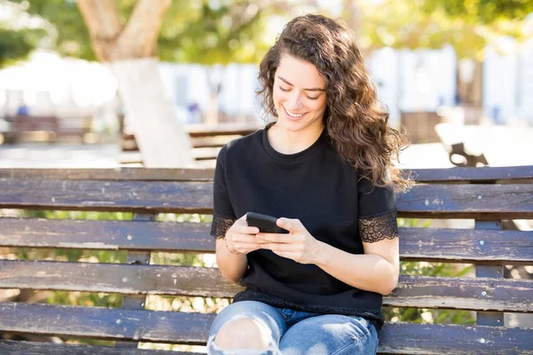 魅力的な若い女性は公園のベンチに座っていると スマート フォンを使用して — ストック写真