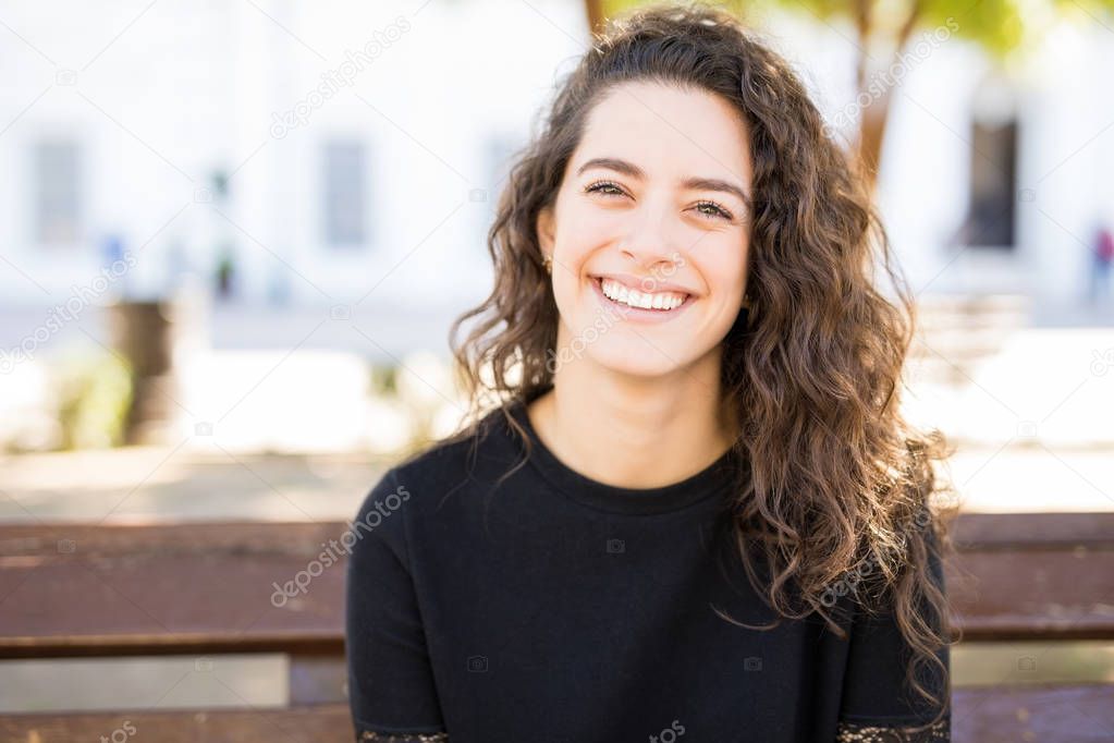 oung brunette smiling while sitting on a bench outside and making an eye contact