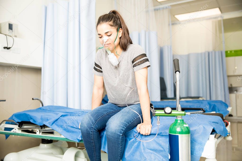 Young latin woman sitting on hospital bed with oxygen mask on getting nebulizer respiratory therapy