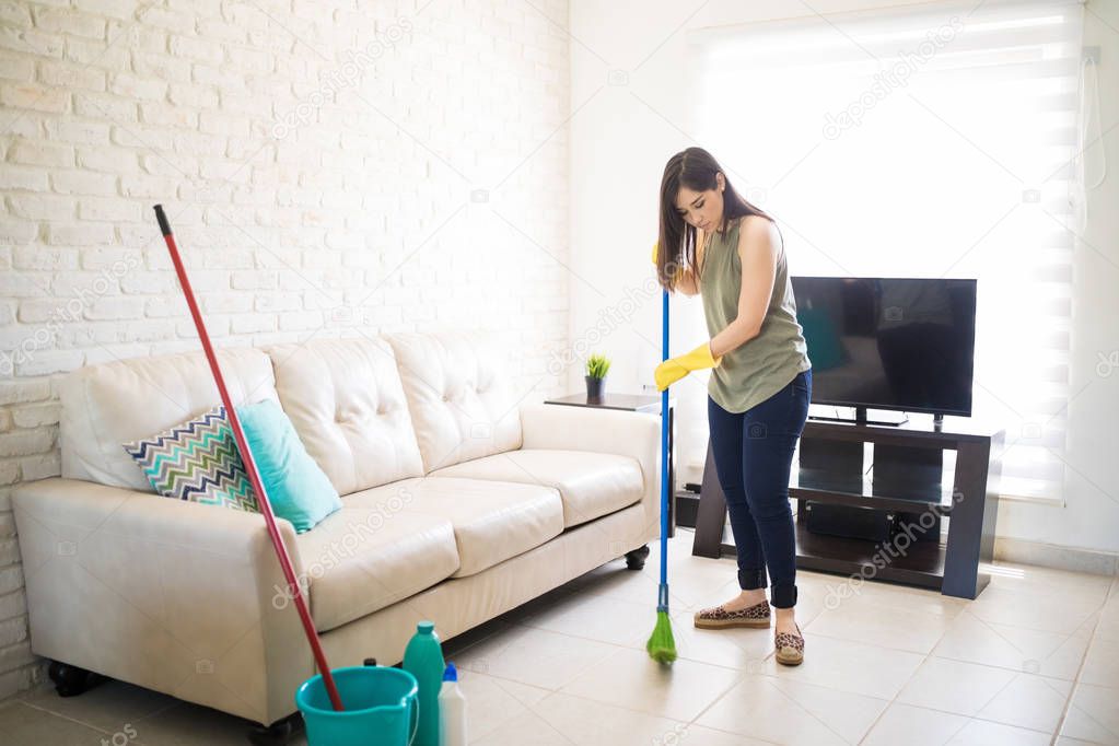 Young maid cleaning house with broom with bucket and disinfectant kept aside near sofa