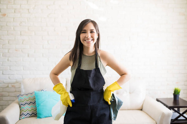 Portrait of a young happy woman wearing black apron and yellow gloves standing holding spray and rug in living room 