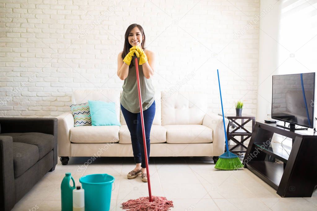 Young woman wearing casuals and yellow rubber gloves cleaning floor while keeping chin on mop