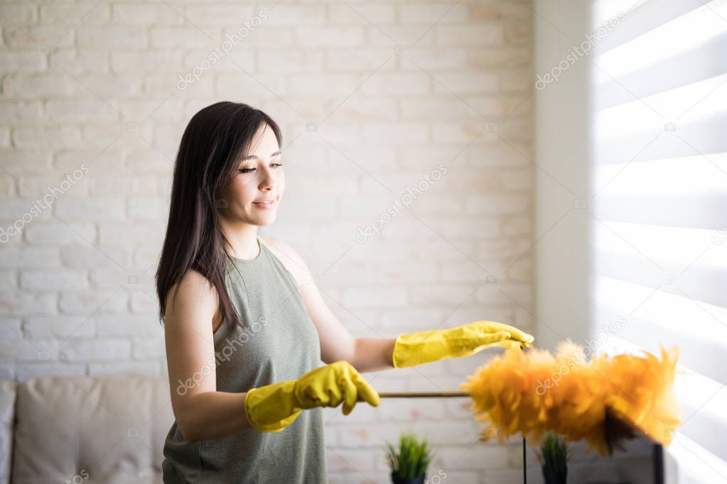 Young woman dusting television top wearing yellow gloves with orange duster