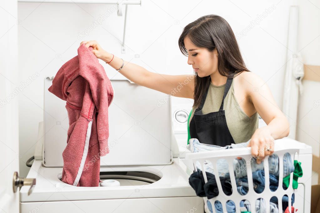 Working woman adding clothes to machine for wash holding white laundry basket