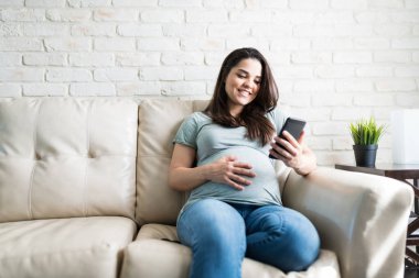 Güzel görünümlü bir kadın oturma odasında elleri karnında otururken akıllı telefon kullanıyor.