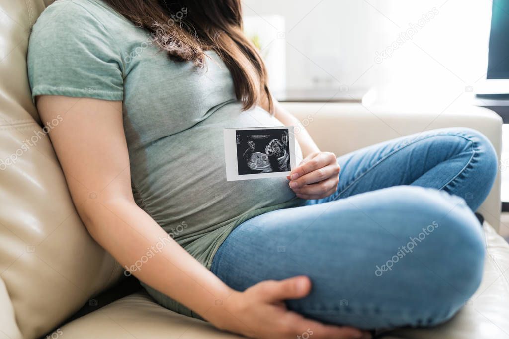 Low section of expectant woman sitting with ultrasound scan on sofa in living room