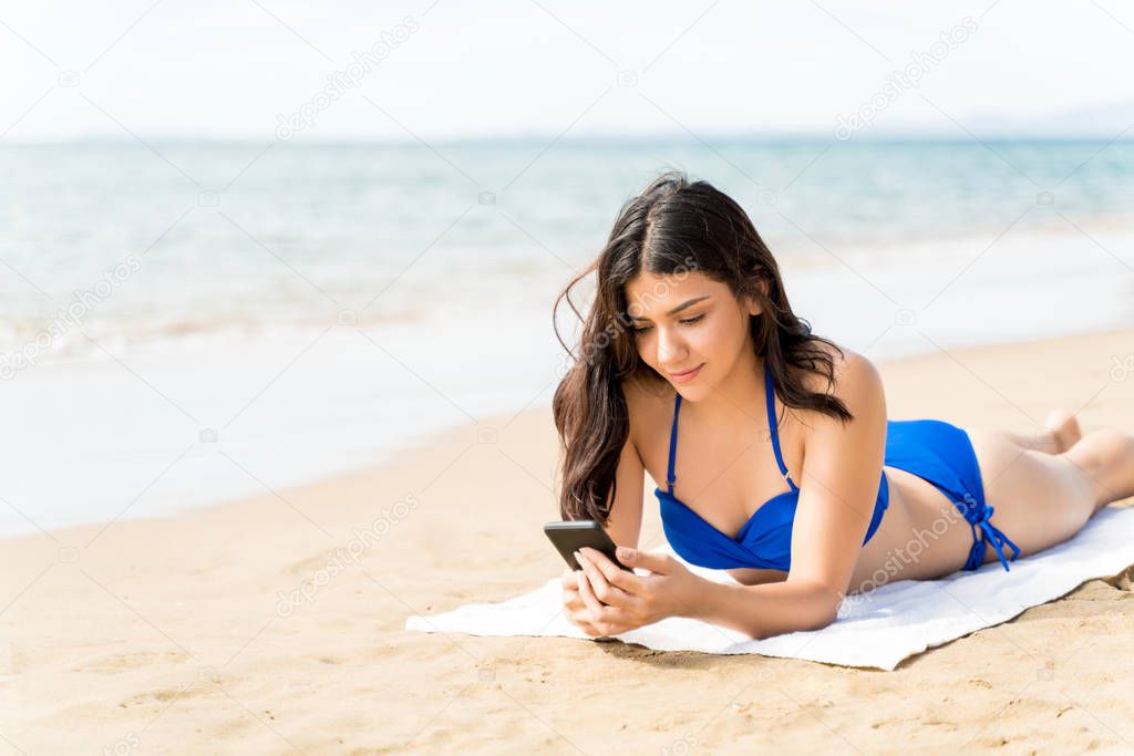 Woman wearing bikini using smartphone while lying on beach towel at sea shore