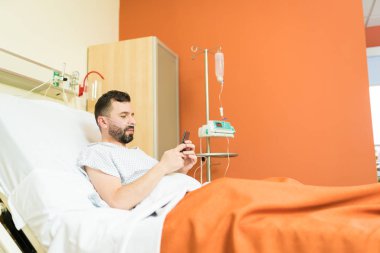 Hastanedeki yatakta dinlenirken akıllı telefondan mesajlaşan yetişkin bir erkek hasta.