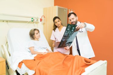 Male and female doctors showing diagnosis of xray to senior patient lying on bed in hospital clipart