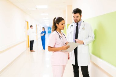 Erkek ve bayan meslektaşlar panoda raporları tartışırken hastane arka planında duran ekip...