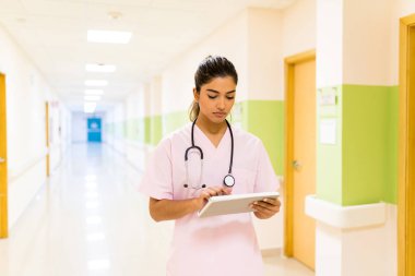 Steteskoplu ciddi bir kadın doktor hastanenin koridorunda yürürken dijital tablet raporunu analiz ediyor.