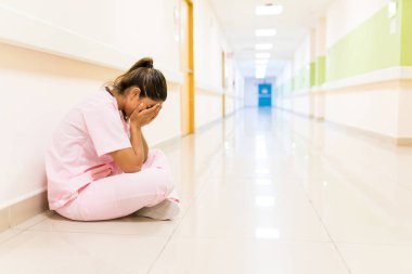 Stressed young nurse covering face while sitting on floor in corridor at hospital clipart