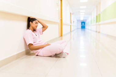 Bitkin düşmüş genç Latin hemşire hastanenin koridorunda duvarın karşısında oturuyor.