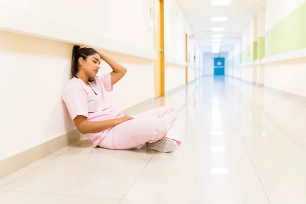 Exhausted young Latin nurse sitting on floor against wall in corridor at hospital