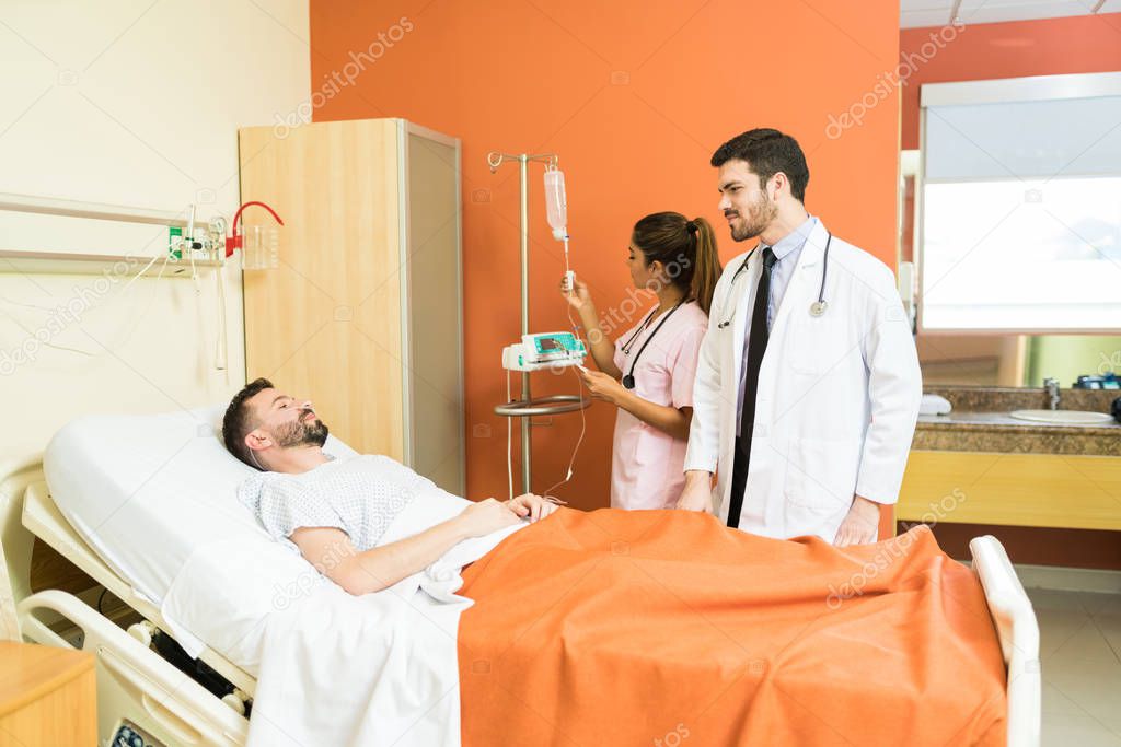 Male doctor talking to patient lying on hospital bed with female healthcare worker adjusting IV drip flow rate