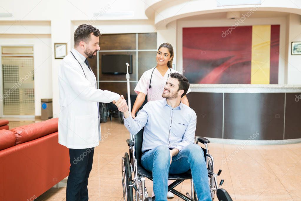Hispanic man smiling on wheelchair while shaking hands with healthcare workers standing at hospital lobby 