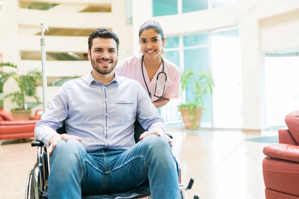 Smiling Latin orderly with mid adult man sitting on wheelchair at lobby in hospital