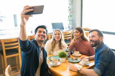 Gülümseyen adam kafeteryada kahve ve tatlının tadını çıkarırken arkadaşlarıyla akıllı telefondan selfie çekiyor.