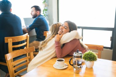 Kafedeki kahve masasında arka planda erkekler otururken kucaklaşan kadın arkadaşlar