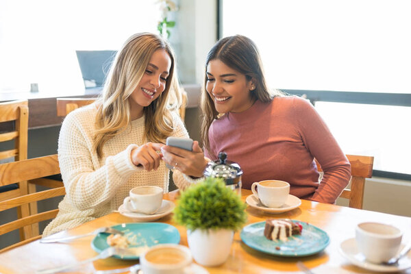 Caucasian woman showing mobile phone to friend while having coffee at table in cafe