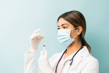 Stüdyoda dikilirken maske takan genç bir doktor şırıngayı kontrol ediyor.