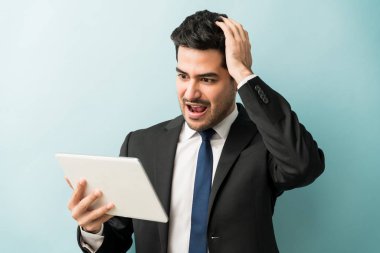 Shocked male professional with hand on head while looking at digital tablet against blue background