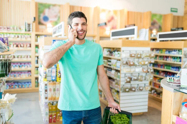 在杂货店拿着菜篮 一边用智能手机笑着的年轻人 — 图库照片