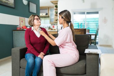 Female caregiver examining elderly woman sitting on sofa at home