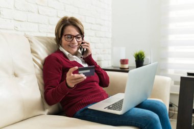 Evdeki kanepede otururken internetten alışveriş yapan gülümseyen emekli kadın.