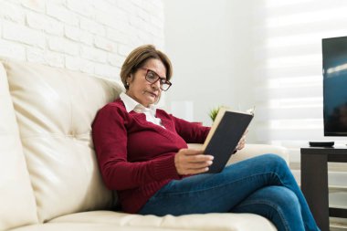 Senior woman reading book while sitting on sofa in living room at home